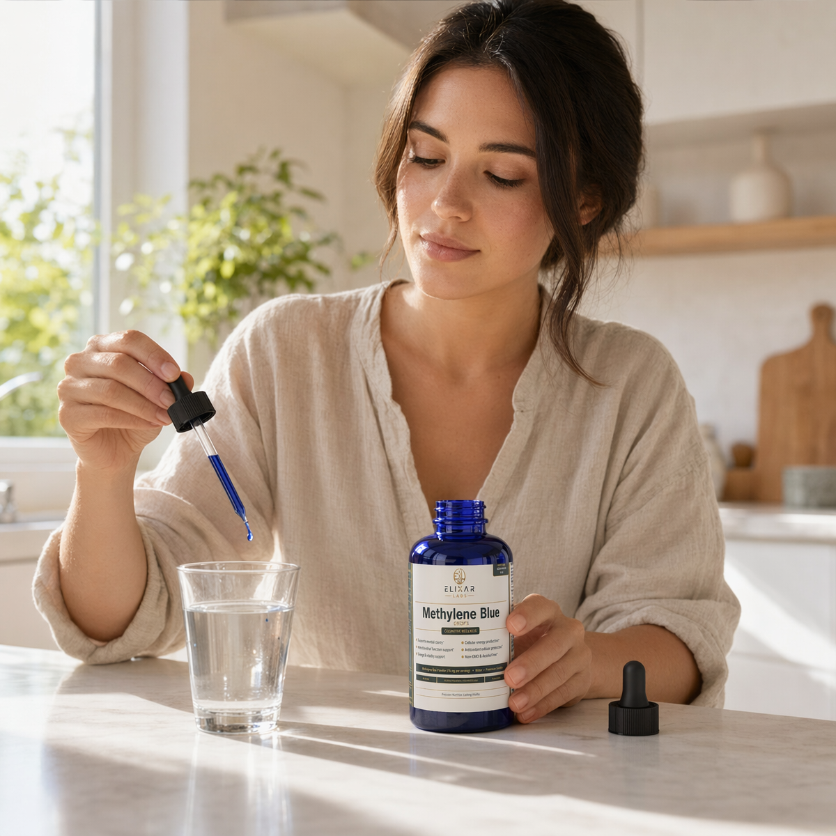 A woman in a beige linen top stands at a kitchen counter holding a glass of water and a blue dropper above a bottle labeled Methylone Blue. The bottle cap is placed beside it, with soft natural light coming from a window in the background.